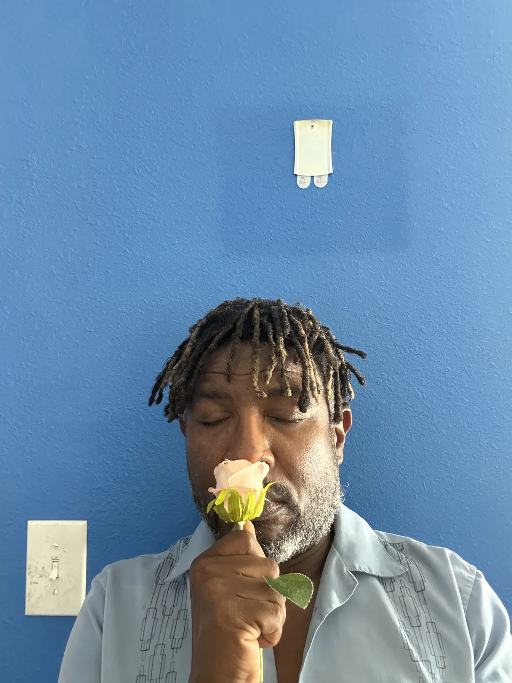 A man with his eyes closed holds a pink rose to his nose against a blue wall